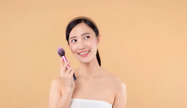 Portrait young happy asian woman with natural make up face holding cosmetic skin powder blusher isolated on beige background. Female apply skincare brush treatment. beauty product cosmetology concept.