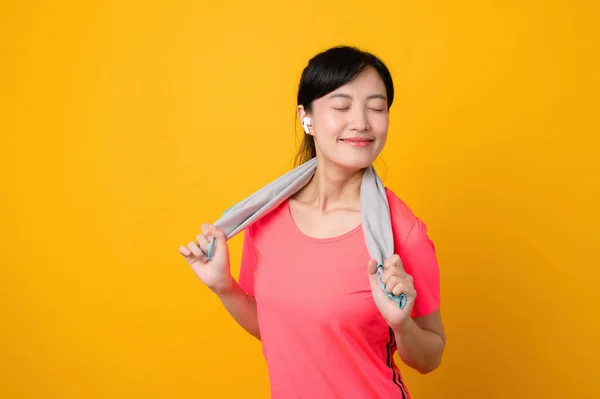 Portrait asian young sports fitness woman happy smile wearing pink sportswear and face towel doing exercise training workout against yellow studio background. wellbeing and healthy lifestyle concept.