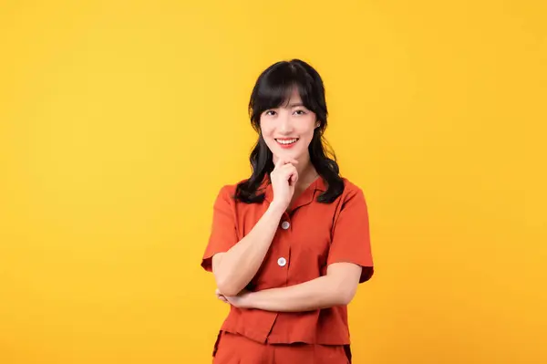 穿着橙色衣服的年轻而体贴的美丽的亚洲女人快乐的微笑 在黄色的工作室背景下表现出思维的姿态 — 图库照片