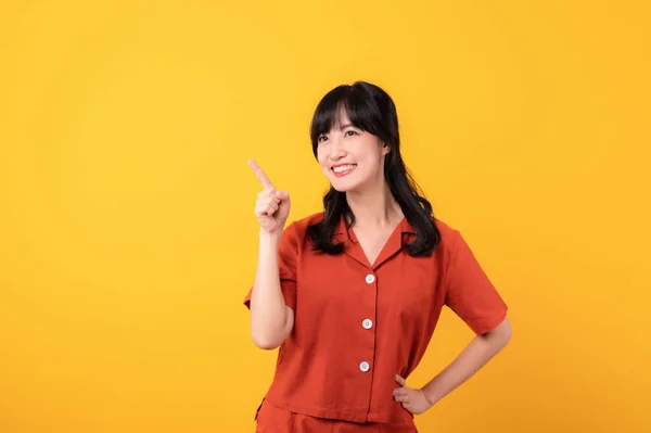 肖像画美しい美しい若いアジアの女性は黄色のスタジオの背景で隔離された自由なスペースに指の手のジェスチャーを指示するオレンジ色の服で幸せな笑顔を示します 素晴らしい製品に注目する — ストック写真