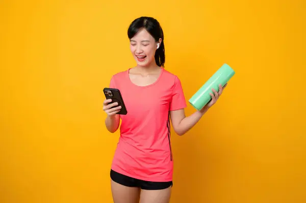 Portrait asian young sports fitness woman happy smile wearing pink sportswear and smartphone doing exercise training workout against yellow studio background. technology wellness lifestyle concept.