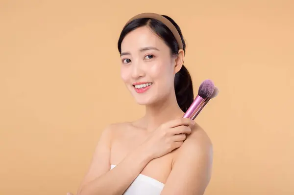 Portrait young happy asian woman with natural make up face holding cosmetic skin powder blusher isolated on beige background. Female apply skincare brush treatment. beauty product cosmetology concept.