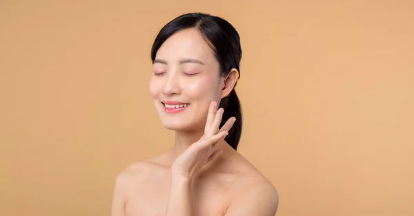 Beautiful asian girl model touching fresh glowing hydrated facial skin on beige background closeup. Beauty face young woman with natural makeup and healthy skin portrait. Skin care concept