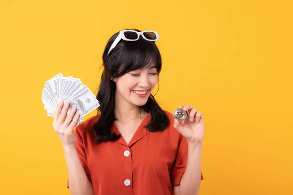 stock image Portrait beautiful young asian woman happy smile dressed in orange clothes holding cash money and crypto bitcoin digital currency celebrate her success isolated on yellow background. Be rich concept.