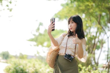 Yeşil park arka planında şapkası, sepeti, cep telefonu ve kamerası olan Asyalı genç bir kadın gezginin portresi. Seyahat yaşam tarzı, dünya turisti ya da Asya yaz turizmi konsepti.