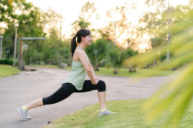 Kadın koşucu. Açık havada koşup eğlenmeden önce yeşil spor kıyafetli sağlıklı bir Asyalı genç kadın parkta kaslarını esnetiyor. Kamu parkındaki spor koşucusu kız. Refah konsept demek.
