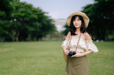 Dokuma şapkası ve sepeti ve yeşil halka açık park arka planında kamerası olan Asyalı genç bir kadın gezginin portresi. Seyahat yaşam tarzı, dünya turisti ya da Asya yaz turizmi konsepti.