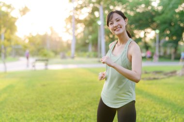 Kadın koşucu. Açık havada koşup eğlenmeden önce yeşil spor kıyafetli sağlıklı bir Asyalı genç kadın parkta kaslarını esnetiyor. Kamu parkındaki spor koşucusu kız. Refah konsept demek.