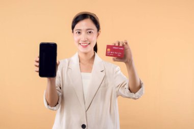 Beyaz ceketli mutlu Asyalı iş kadınının portresi. Elinde akıllı telefon ve kredi kartı bej arka planda duruyor. alışveriş kavramı.