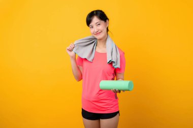 Asyalı genç spor fitness kadını portresi. Pembe spor kıyafetleri ve yüz havlusu giymiş mutlu bir gülümseme. Sarı stüdyo arka planında egzersiz yapıyor. Refah ve sağlıklı yaşam tarzı kavramı.