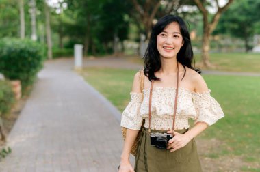 Dokuma şapkası ve sepeti ve yeşil halka açık park arka planında kamerası olan Asyalı genç bir kadın gezginin portresi. Seyahat yaşam tarzı, dünya turisti ya da Asya yaz turizmi konsepti.
