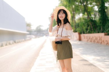 Portrait of young asian woman traveler with weaving hat and basket and a camera standing by the street. Journey trip lifestyle, world travel explorer or Asia summer tourism concept.