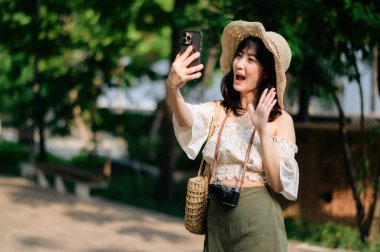 Yeşil park arka planında şapkası, sepeti, cep telefonu ve kamerası olan Asyalı genç bir kadın gezginin portresi. Seyahat yaşam tarzı, dünya turisti ya da Asya yaz turizmi konsepti.