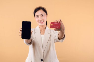 Beyaz ceketli mutlu Asyalı iş kadınının portresi. Elinde akıllı telefon ve kredi kartı bej arka planda duruyor. alışveriş kavramı.