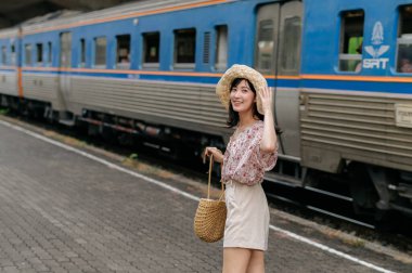 Elinde sepet olan Asyalı genç bir kadın gezgin, arkasını dönüp arkasına bile bakmadan trene gülümsüyor. Seyahat yaşam tarzı, dünya turisti ya da Asya yaz turizmi konsepti.