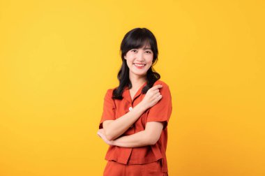 Portre genç, düşünceli güzel Asyalı mutlu bir gülümseme turuncu giysiler içinde sarı stüdyo arka planında izole edilmiş bir jest gösteriyor..