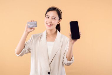 Beyaz ceketli mutlu Asyalı iş kadınının portresi. Elinde akıllı telefon ve kredi kartı bej arka planda duruyor. alışveriş kavramı.