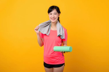 Asyalı genç spor fitness kadını portresi. Pembe spor kıyafetleri ve yüz havlusu giymiş mutlu bir gülümseme. Sarı stüdyo arka planında egzersiz yapıyor. Refah ve sağlıklı yaşam tarzı kavramı.