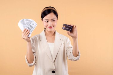 Beyaz ceketli mutlu Asyalı iş kadınının portresi. Elinde nakit para ve kredi kartı var. Bej arka planda duruyor. Milyoner işi, alışveriş konsepti