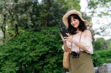 Yeşil park arka planında şapkası, sepeti, cep telefonu ve kamerası olan Asyalı genç bir kadın gezginin portresi. Seyahat yaşam tarzı, dünya turisti ya da Asya yaz turizmi konsepti.