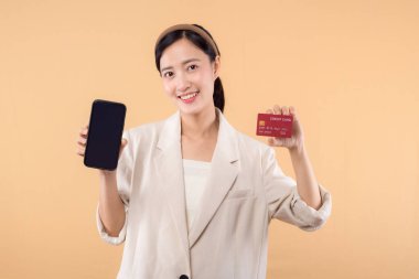 Beyaz ceketli mutlu Asyalı iş kadınının portresi. Elinde akıllı telefon ve kredi kartı bej arka planda duruyor. alışveriş kavramı.