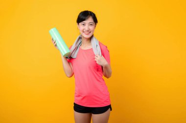Asyalı genç spor fitness kadını portresi. Pembe spor kıyafetleri ve yüz havlusu giymiş mutlu bir gülümseme. Sarı stüdyo arka planında egzersiz yapıyor. Refah ve sağlıklı yaşam tarzı kavramı.