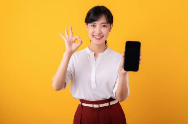 Genç, güzel Asyalı girişimci kadının portresi beyaz gömlek ve kırmızı bitkiler giyerek mutlu bir gülümseme takınıyor ve sarı arka plandaki akıllı telefon izolasyonuna el hareketi yapıyor. Yeni uygulama kavramı sunuluyor