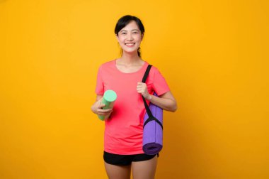 Asyalı genç spor fitness kadını portresi. Pembe spor kıyafetleri ve yoga minderi giymiş mutlu bir gülümseme. Sarı stüdyo arka planında egzersiz yapıyor. Sağlıklı sağlık yaşam tarzı kavramı.