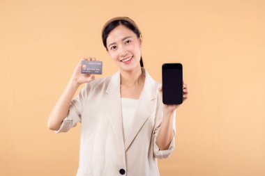 Beyaz ceketli mutlu Asyalı iş kadınının portresi. Elinde akıllı telefon ve kredi kartı bej arka planda duruyor. alışveriş kavramı.