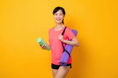 Asyalı genç spor fitness kadını portresi. Pembe spor kıyafetleri ve yoga minderi giymiş mutlu bir gülümseme. Sarı stüdyo arka planında egzersiz yapıyor. Sağlıklı sağlık yaşam tarzı kavramı.
