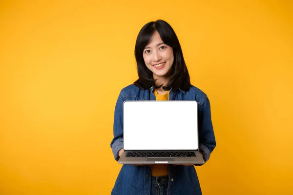 portrait happy young woman wearing yellow t-shirt and denim shirt holding laptop and point finger to screen isolated on yellow studio background. business technology application communication concept.