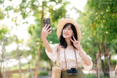 Yeşil park arka planında şapkası, sepeti, cep telefonu ve kamerası olan Asyalı genç bir kadın gezginin portresi. Seyahat yaşam tarzı, dünya turisti ya da Asya yaz turizmi konsepti.