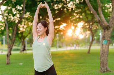 Kadın koşucu. Açık havada koşup eğlenmeden önce yeşil spor kıyafetli sağlıklı bir Asyalı genç kadın parkta kaslarını esnetiyor. Kamu parkındaki spor koşucusu kız. Refah konsept demek.