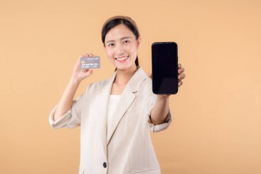 Beyaz ceketli mutlu Asyalı iş kadınının portresi. Elinde akıllı telefon ve kredi kartı bej arka planda duruyor. alışveriş kavramı.