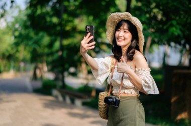 Yeşil park arka planında şapkası, sepeti, cep telefonu ve kamerası olan Asyalı genç bir kadın gezginin portresi. Seyahat yaşam tarzı, dünya turisti ya da Asya yaz turizmi konsepti.
