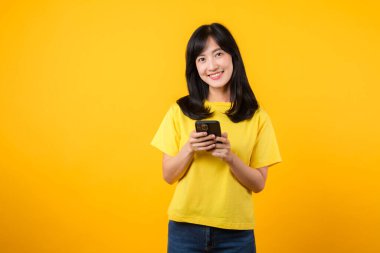 Teknolojinin mutluluğunu ve bağlantısını neşeli bir portre ile tecrübe edin. Sarı tişört ve kot pantolon giyen Asyalı genç kadın akıllı telefon kullanarak gülümsüyor. Teknoloji ve akıllı telefon uygulaması kavramı.