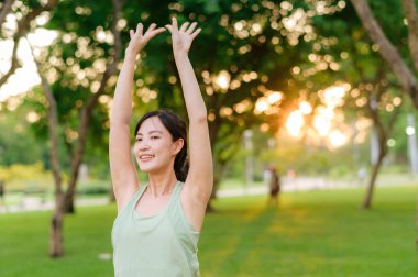 Kadın koşucu. Açık havada koşup eğlenmeden önce yeşil spor kıyafetli sağlıklı bir Asyalı genç kadın parkta kaslarını esnetiyor. Kamu parkındaki spor koşucusu kız. Refah konsept demek.