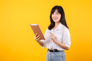 Beyaz gömlek giyen Asyalı genç bir kadın sarı arka planda dijital tableti tutarken parlak bir şekilde gülümsüyor, modern teknoloji kavramlarını benimseyen kendine güvenen bir şirket çalışanını temsil ediyor..