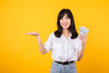 Asya kökenli mutlu genç bir kadın beyaz gömlek ve kot pantolon giyer ve elinde dolar olarak nakit para tutar ve sarı arka planda bedava fotokopi çekilir. Yatırım ve finansal konsept.