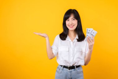 Asya kökenli mutlu genç bir kadın beyaz gömlek ve kot pantolon giyer ve elinde dolar olarak nakit para tutar ve sarı arka planda bedava fotokopi çekilir. Yatırım ve finansal konsept.