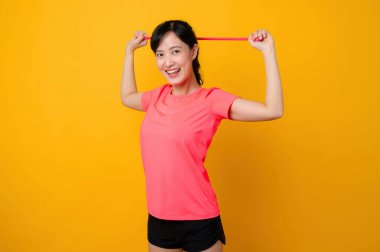 Asyalı genç spor formalı kadın, pembe spor giyerek ve direnç bandını gererek sarı arka planda egzersiz yaparak mutlu oluyor. Sağlıklı yaşam tarzı kavramı.