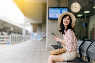 Cep telefonu kullanıp tren istasyonunda bekleyen sepetli Asyalı genç bir kadın gezgin. Seyahat yaşam tarzı, dünya turisti ya da Asya yaz turizmi konsepti.