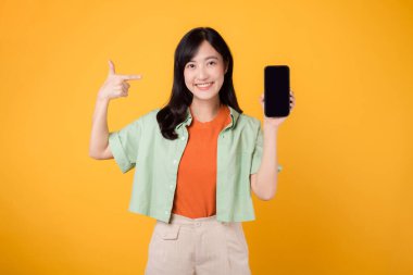 Yeşil ve turuncu gömlek giyen 30 'lu yaşlardaki neşeli Asyalı kadın. Akıllı telefonu işaret ediyor, bağlantıyı ve dijital güçlenmeyi sembolize ediyor. Teknoloji ve modern yaşam tarzı canlı sarı bir arkaplanda..