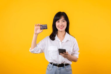 Beyaz gömlekli ve kot pantolonlu genç Asyalı bir kadının portresi. Sarı arka planda izole edilmiş akıllı telefondan online alışveriş yapmak için kredi kartı taşıyor. alışveriş çevrimiçi kavramı.