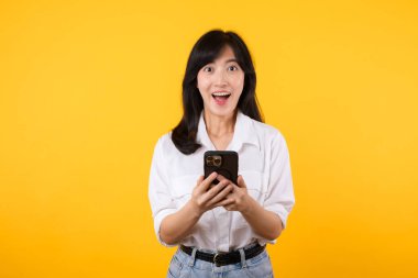 Asyalı genç bir kadının cep telefonunu olumlu bir ifadeyle kullanmasına şaşırdım. Geniş bir şekilde gülümsüyor, beyaz gömlek giyiyor ve sarı arka planda tek başına duruyor. Mutlu, mutlu, mutlu kadın başarıya sevinir..