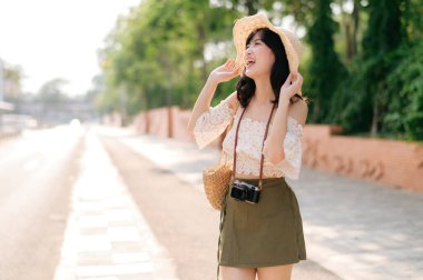 Portrait of young asian woman traveler with weaving hat and basket and a camera standing by the street. Journey trip lifestyle, world travel explorer or Asia summer tourism concept.