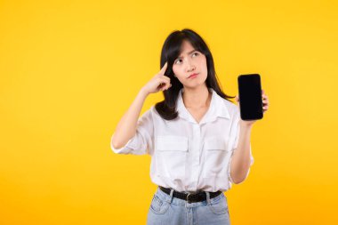 Şüpheli yüzü olan güzel Asyalı bir kadının portresi beyaz gömlek ve akıllı telefon eşliğinde kot pantolon giymesi sarı stüdyo arka planında izole edilmiş. Sahte online haber konseptiyle hayal kırıklığına uğradım..