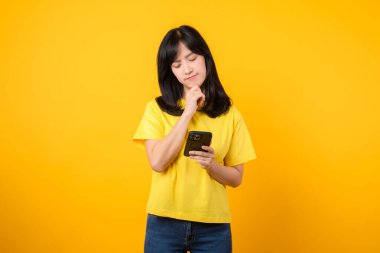 Etkileyici bir portre ile duyguların deneyim aralığı. Sarı tişört ve kot pantolon giyen Asyalı genç kadın akıllı telefon kullanırken şüpheli bir ifade sergiliyor. Çeşitli duygular ve teknolojik kullanım.