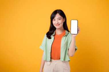 Portre mutlu gülümseme turuncu tişört giyen 30 'lu yaşlardaki Asyalı kadın sarı stüdyo arka planında izole edilmiş akıllı telefon görüntüleme cihazı gösteriyor. reklam yeni teknoloji mobil uygulama kavramı.