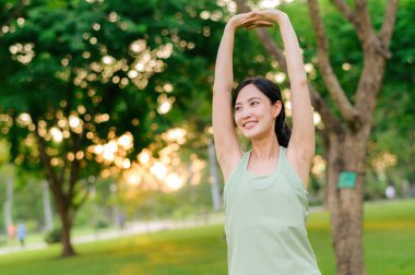 Kadın koşucu. Açık havada koşup eğlenmeden önce yeşil spor kıyafetli sağlıklı bir Asyalı genç kadın parkta kaslarını esnetiyor. Kamu parkındaki spor koşucusu kız. Refah konsept demek.
